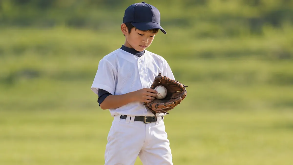 守備位置でグラブのボールを見てひと息つく少年のイメージ（生成AIによる画像）